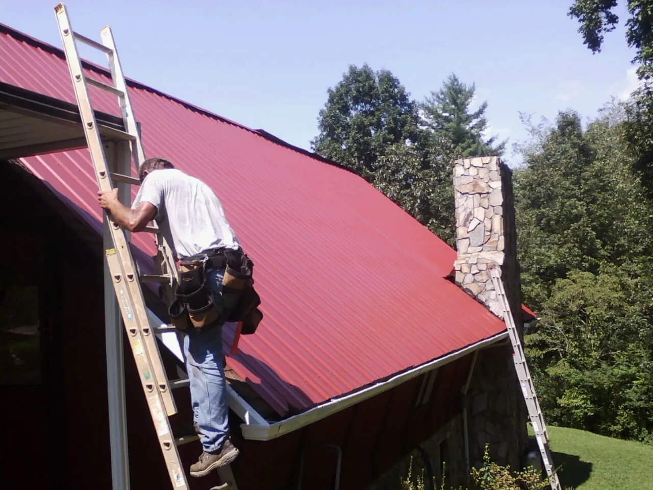 Roofer on a ladder beside a red metal roof and stone chimney — Soffit & Fascia Repair by Roofing Brothers in Slingerlands
